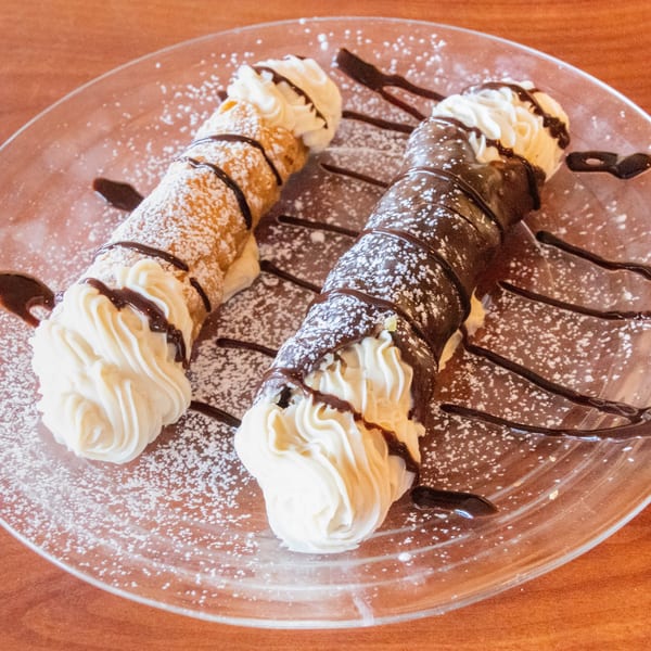 Two cannolis, one plain and one chocolate-dipped, topped with whipped cream and chocolate drizzle.