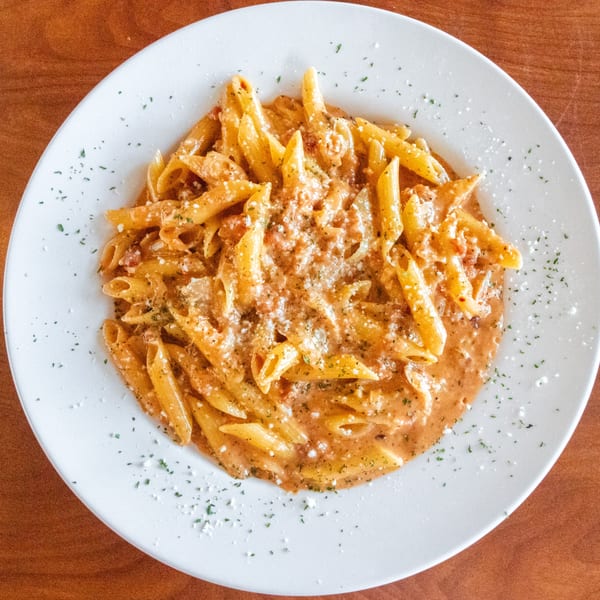 A white plate filled with penne pasta in a creamy red sauce, topped with Parmesan and herbs.