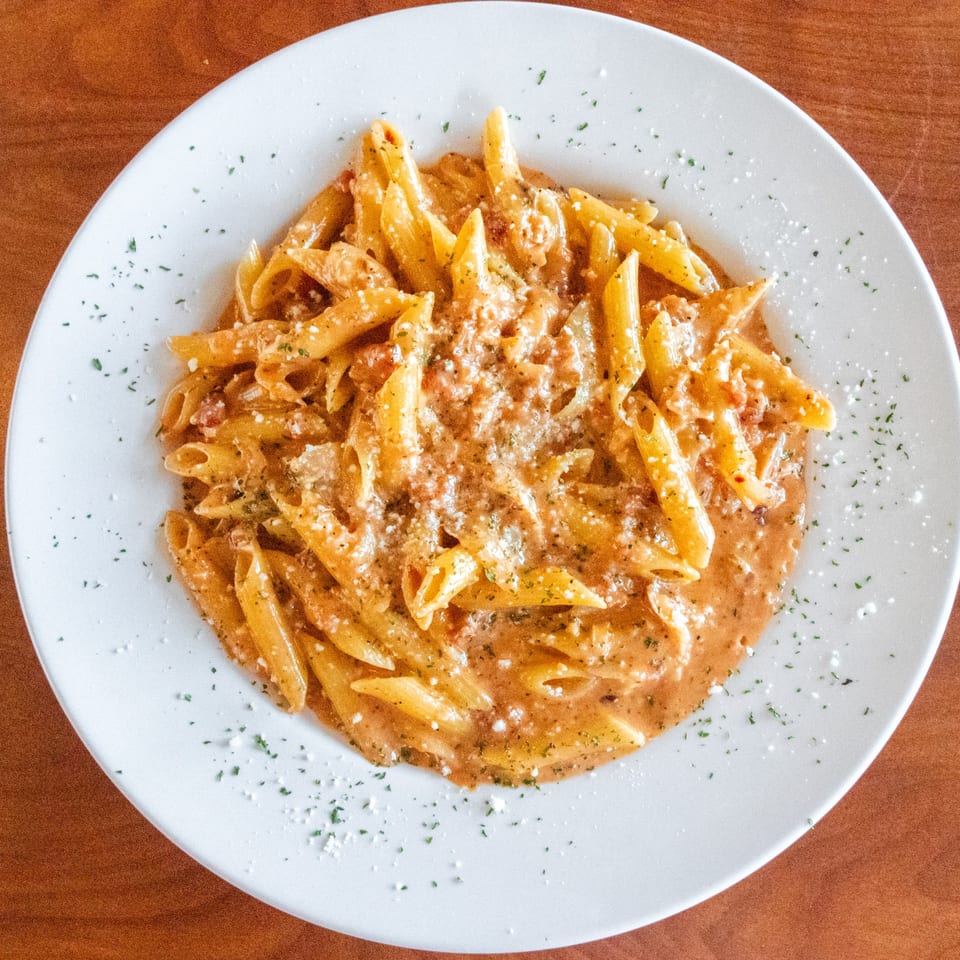 A white plate filled with penne pasta in a creamy red sauce, topped with Parmesan and herbs.